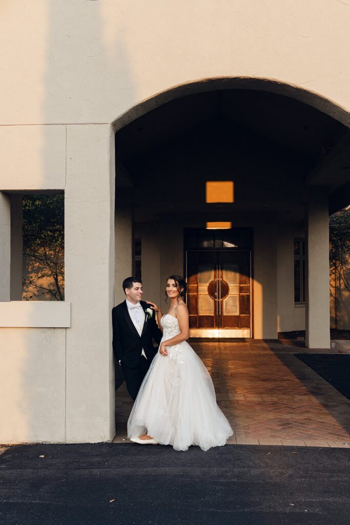 Ana and Tyler pose beneath RiverStone's arched covered entrance, with warm evening light reflecting on the wooden double doors behind them.