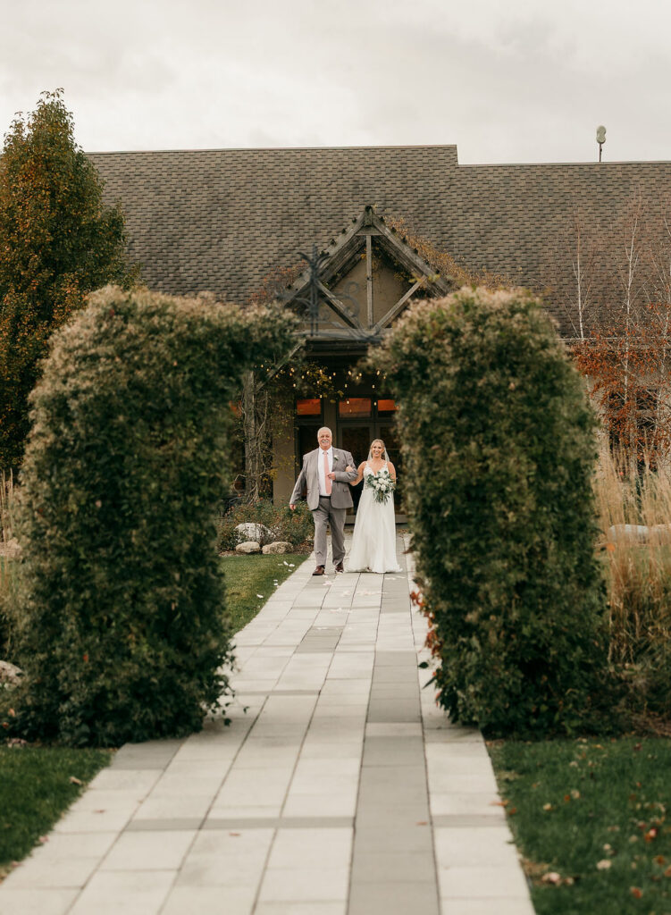 Kristy walks arm-in-arm with her father down RiverStone's stone garden aisle, framed by tall manicured hedges.