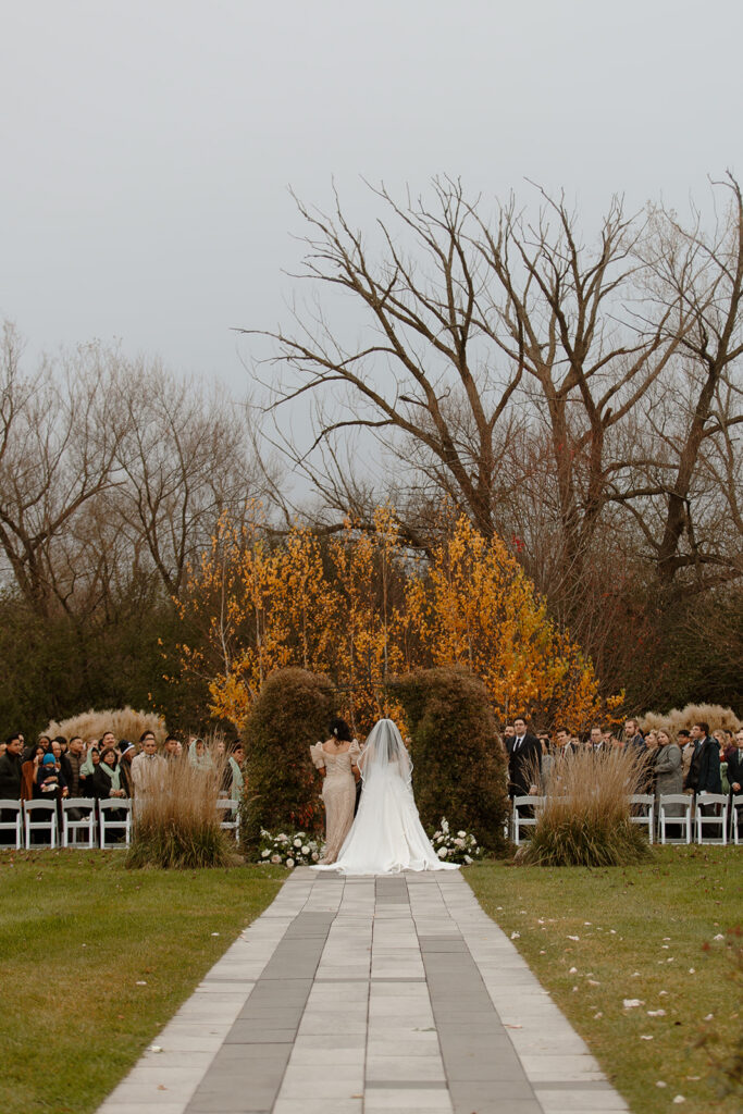 Wide view of Mariah and Nathan's outdoor wedding ceremony at RiverStone, with a stone aisle, seated guests, and autumn trees.