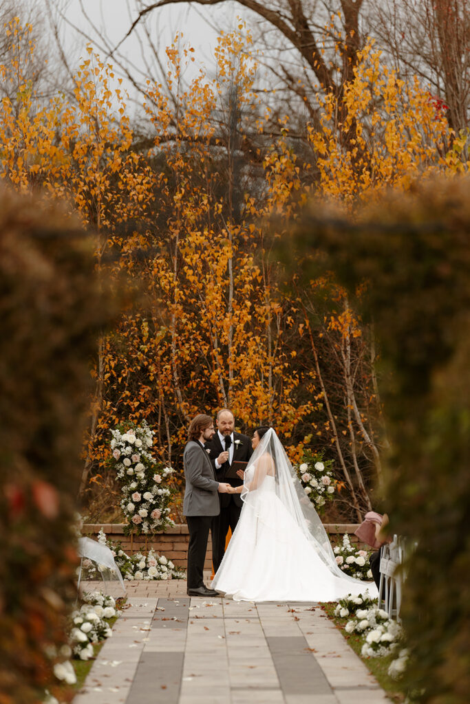 Mariah and Nathan exchange vows at RiverStone's outdoor garden altar, surrounded by floral arrangements and fall foliage.