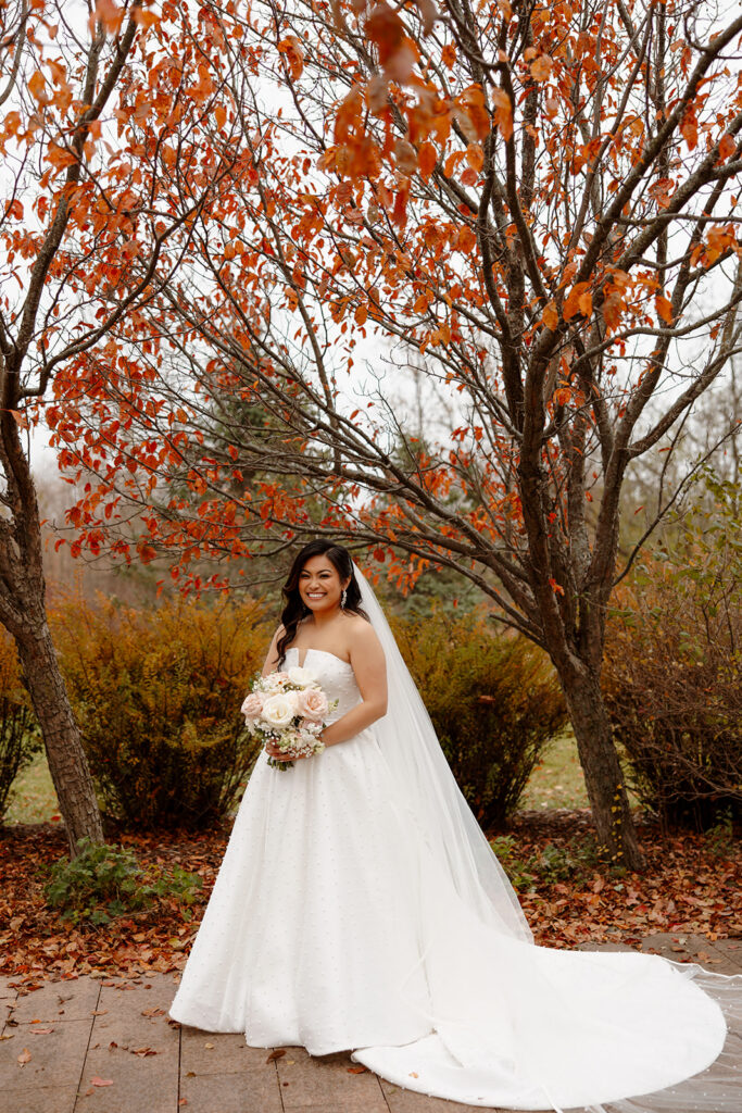 Mariah holds her blush and ivory wedding bouquet beneath vibrant orange autumn leaves in RiverStone's gardens.
