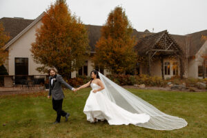 Mariah and Nathan walk together across RiverStone's green lawn, with the wedding venue and golden fall trees in the background.