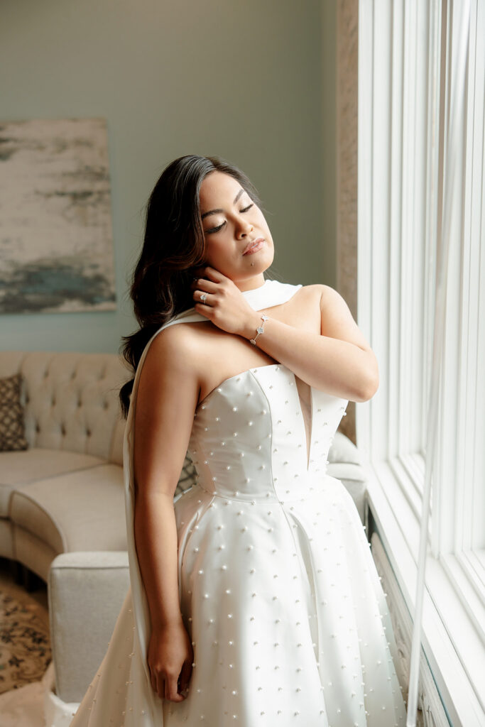 Bride Mariah poses beside the tall windows of RiverStone's bridal suite, with soft natural light illuminating her wedding gown.