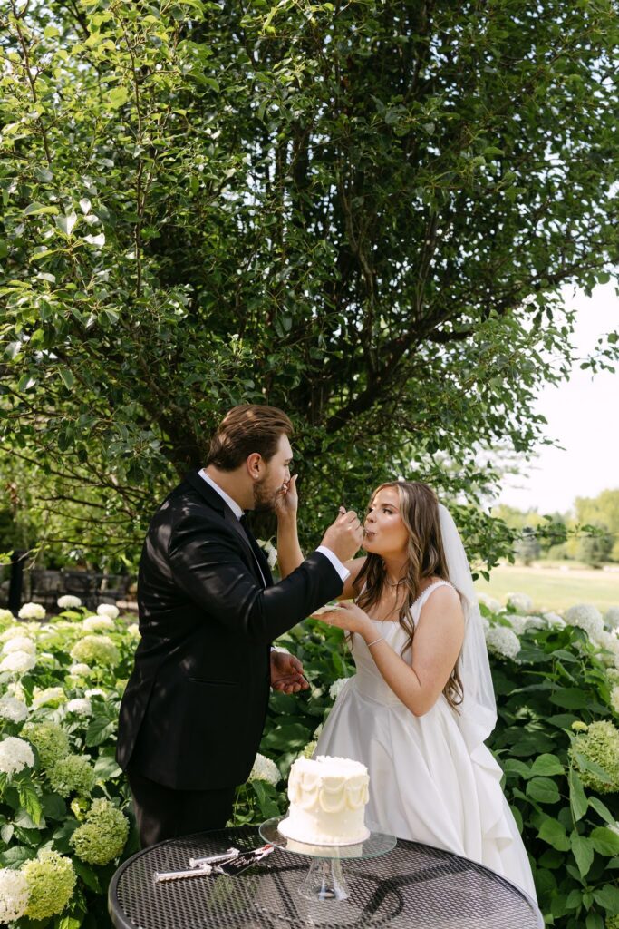 Sophie and Tyson share a bite of wedding cake outdoors beside blooming white hydrangeas at RiverStone.