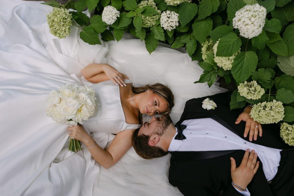 Sophie and Tyson lie together on the bride's flowing white gown surrounded by blooming hydrangeas at RiverStone.