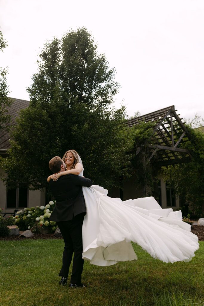 Tyson lifts Sophie into his arms as her wedding dress flows through the air in RiverStone's gardens.