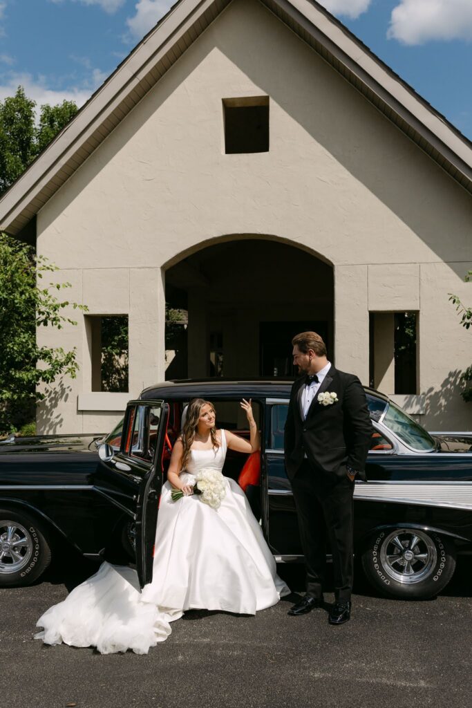 Sophie and Tyson pose beside a black vintage car outside RiverStone during their July wedding.